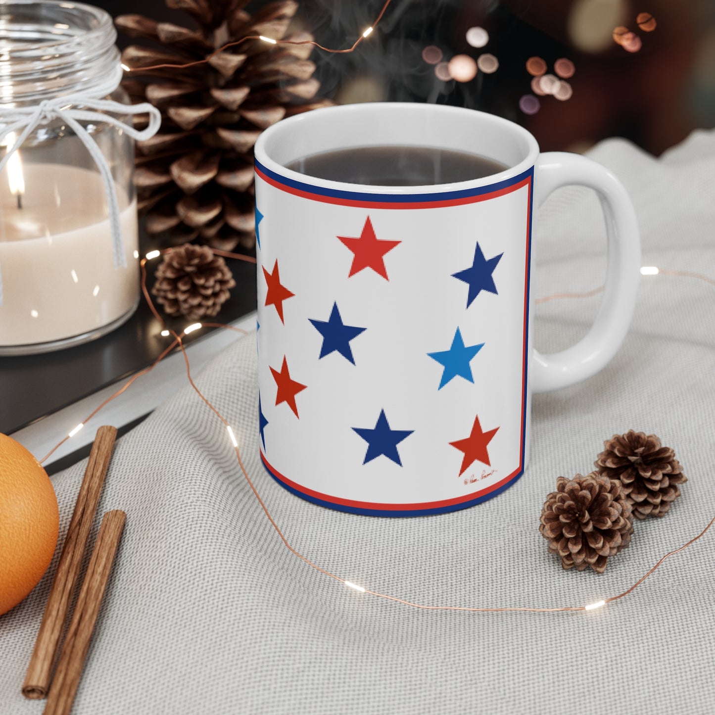 A patriotic Mug with Stars: Blue and Red on White; Ceramic; 11 oz. by Printify sits on a cloth next to pine cones, cinnamon sticks, an orange, and a lit candle in a jar.
