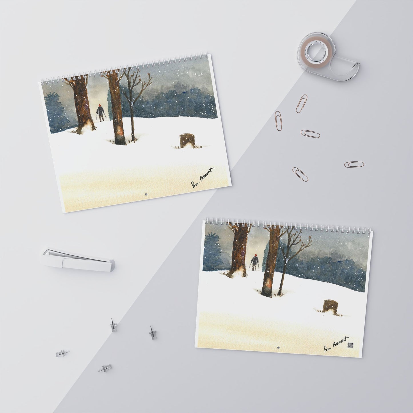 Two identical 2025 Nature-Themed Wall Calendars from Printify, showcasing watercolor art of a winter scene with trees and snow, are elegantly displayed on a two-toned surface. Enhancing the artistic ambiance are surrounding items such as a tape dispenser, paper clips, a stapler, and thumbtacks.