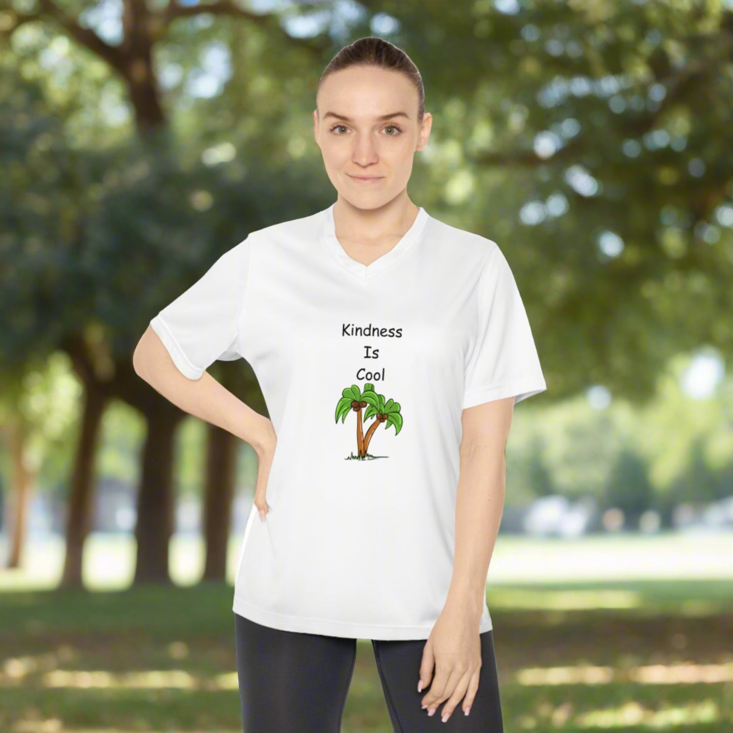 A person stands outdoors wearing a Printify Women's Kindness-Is-Cool V-neck T-shirt, made of moisture-wicking polyester with UV 40+ protection, featuring the text "Kindness Is Cool" printed above an image of a palm tree from Team 365.