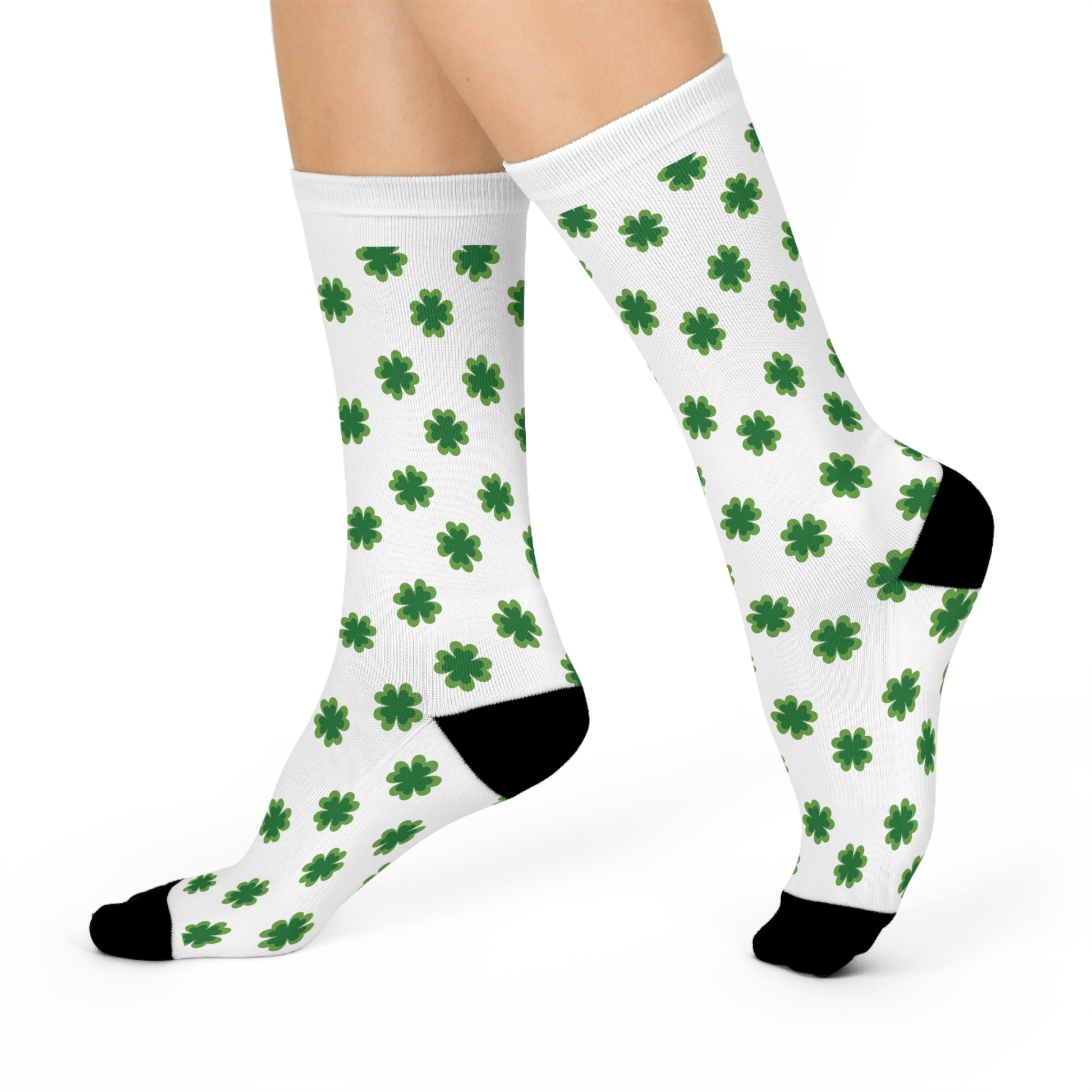 A person wears Printify's Shamrock-Pattern Cushioned Crew-Socks: Unisex | St. Patrick's Day—white socks with black toes and heels, featuring repeating green four-leaf clovers, displaying both feet.
