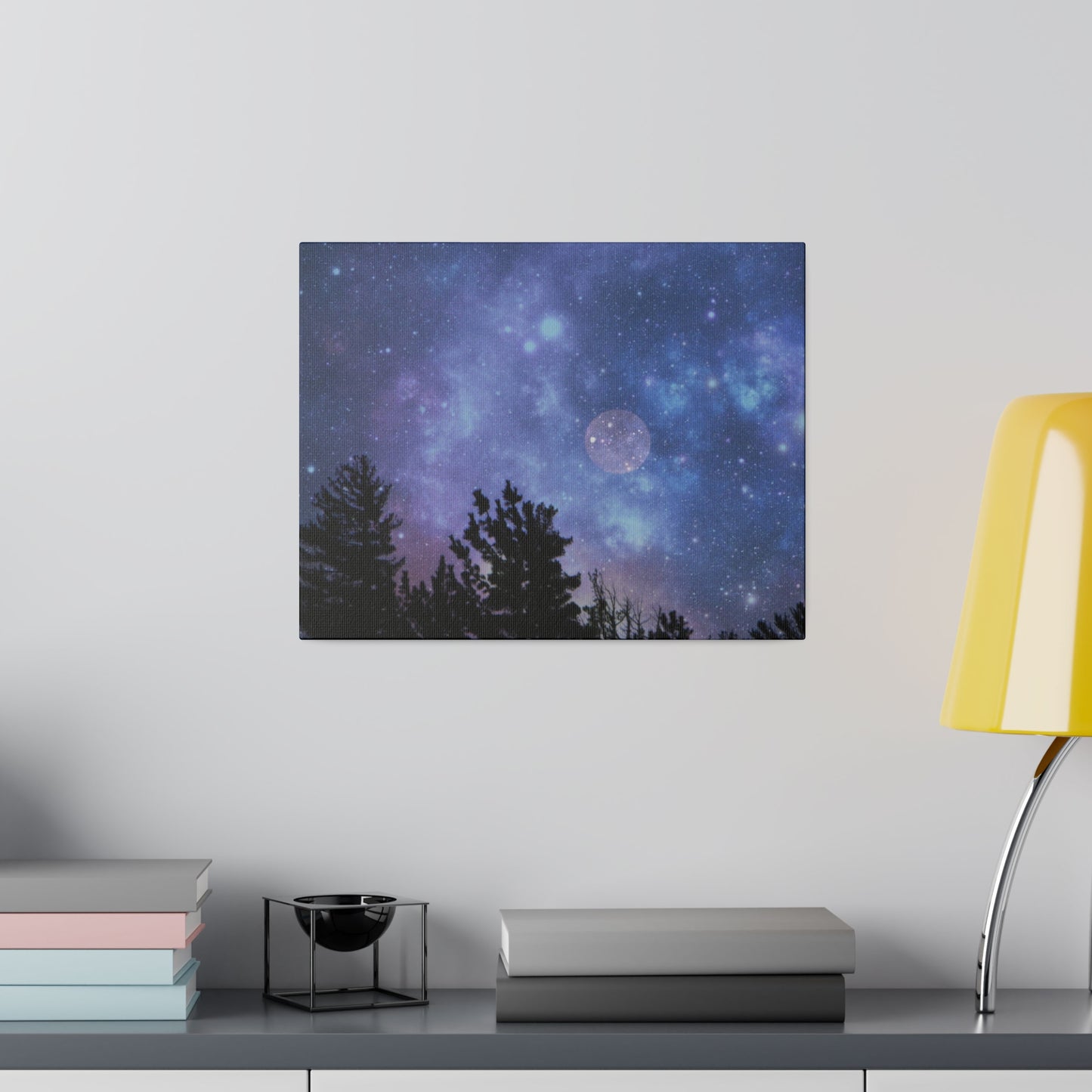 A cosmic wall art featuring a starry night sky and silhouette of trees above a stack of books, portrayed on a Printify Blue-Moon Matte Canvas next to a yellow lamp.