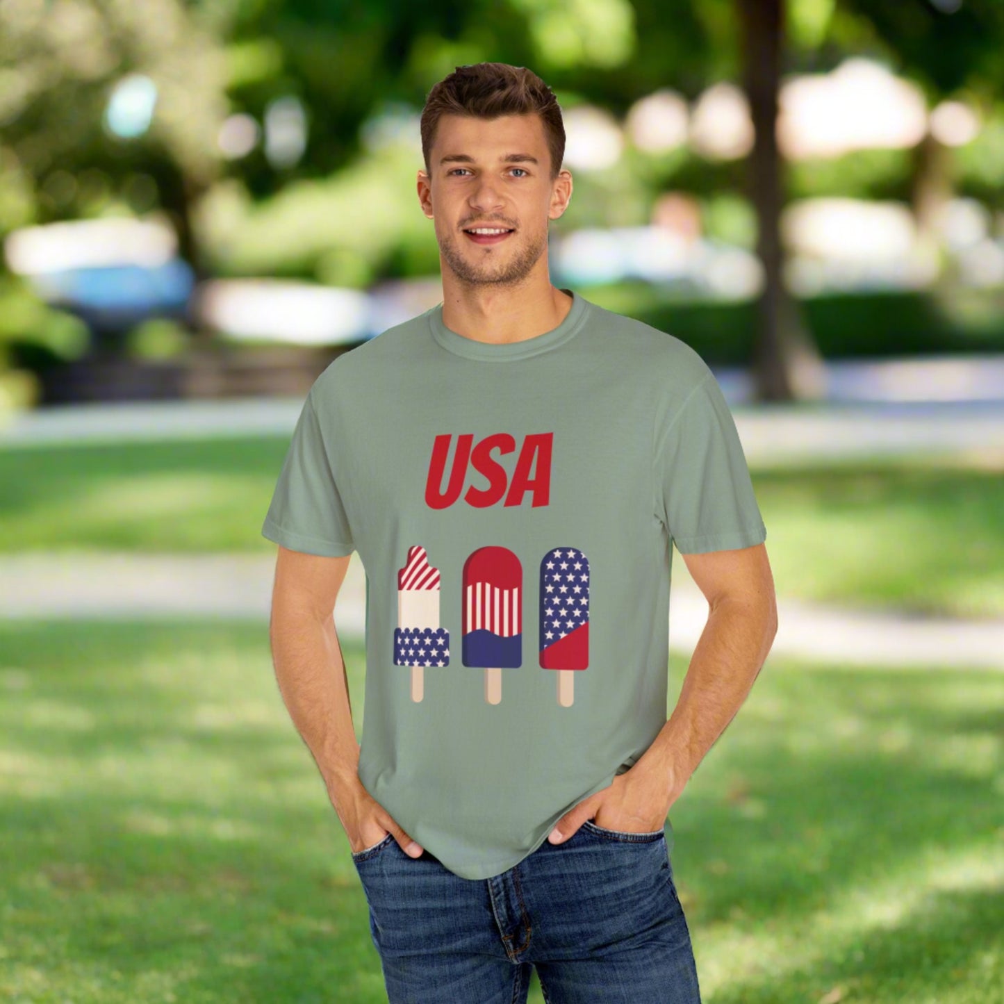 A man stands outdoors wearing a light green Unisex USA T-shirt: 3 colors; Cotton; Comfort Colors 1717 with "USA" and three popsicles in American flag colors printed on it. Made from 100% ring-spun cotton by Printify, the T-shirt complements his casual look. He has short hair and has his hands in his pockets. Trees and shrubs are in the background.