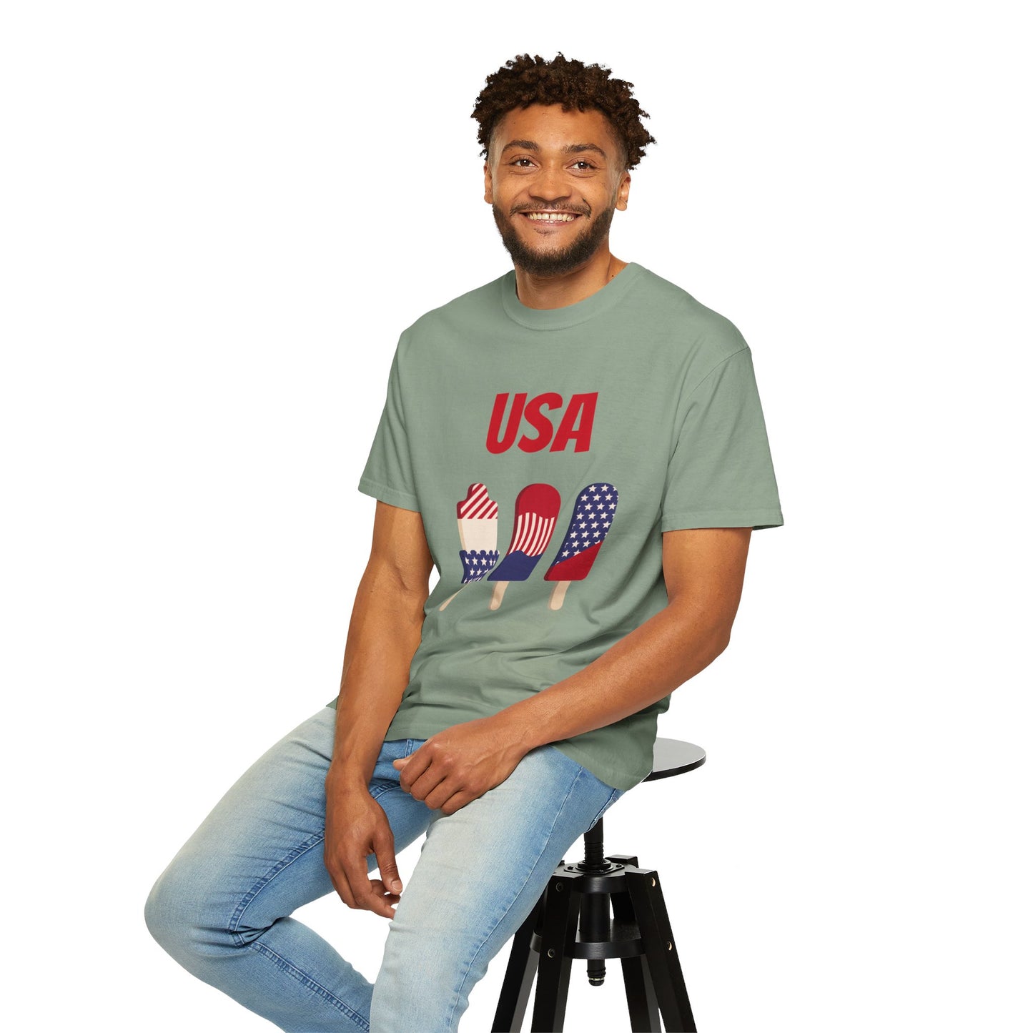 A man with a beard wearing a green Unisex USA T-shirt: 3 colors; Cotton; Comfort Colors 1717 from Printify, featuring popsicles in American flag designs, sits on a stool, smiling.