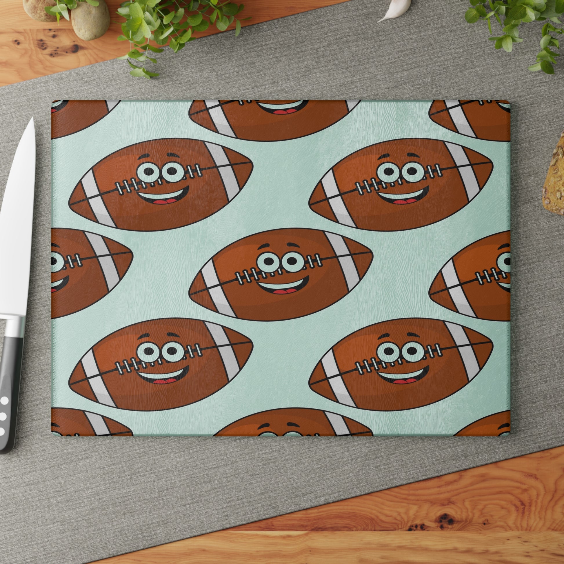 The Printify Playful Football-Faces Cutting Board (glass, 2 sizes) features a fun pattern of smiling footballs—an ideal kitchen decor piece and perfect for sports fans. Shown on a gray mat with a knife and greenery.