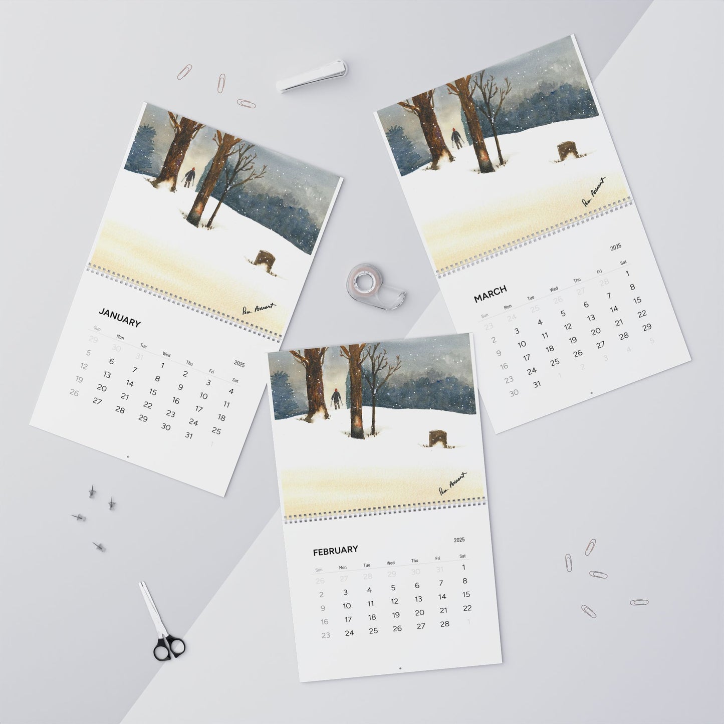 On a desk scattered with office supplies such as scissors, paperclips, and tape, lay three winter-themed pages for January, February, and March. Each page features a stunning watercolor print from the Printify 2025 Nature-Themed Wall Calendar available in two sizes.