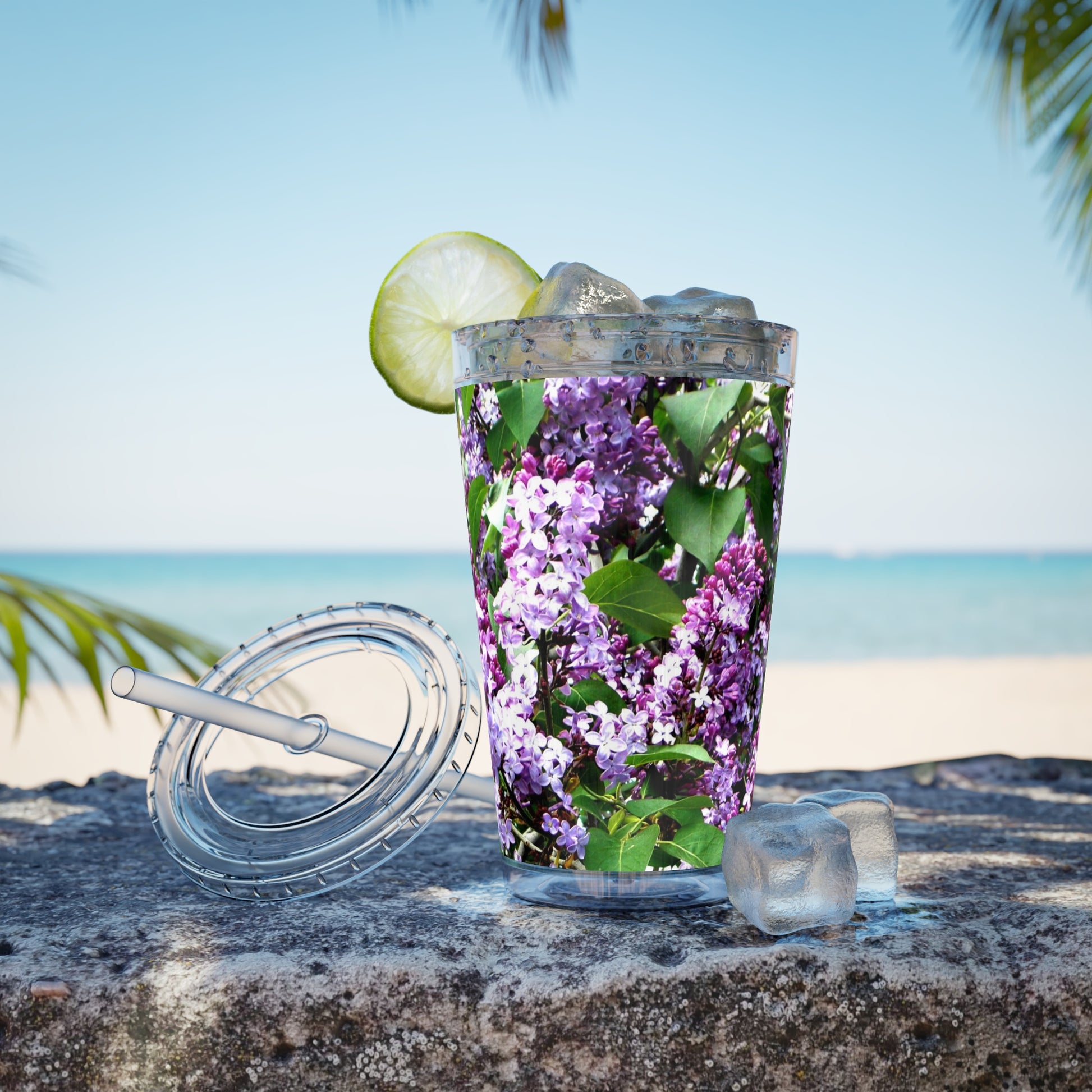 A Purple Lilacs Tumbler from Printify with lilacs and lime on the beach.
