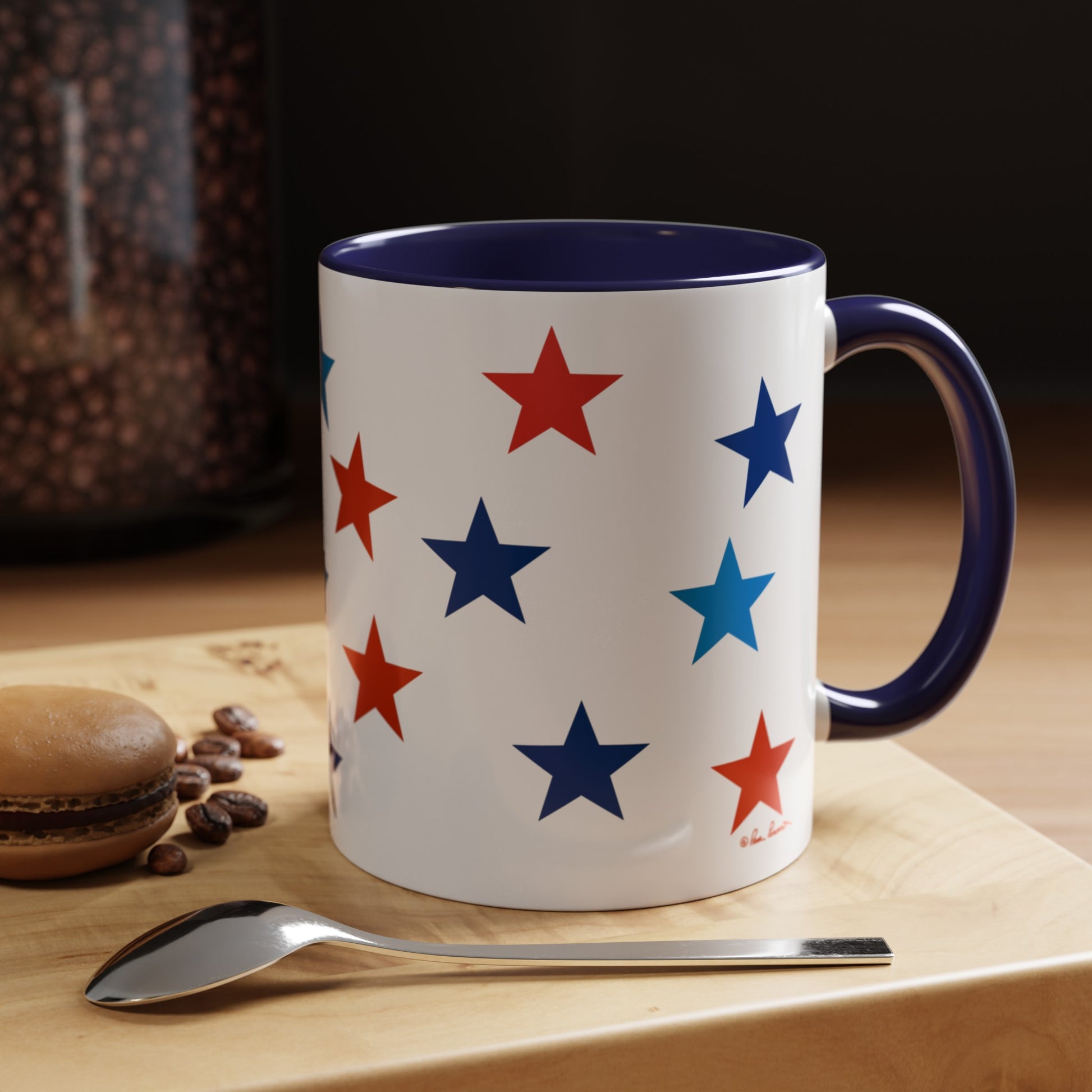 A Printify Patriotic Coffee Mug: 11 oz.; 2 Accent colors; Ceramic adorned with red and blue stars sits on a table next to a spoon and a macaron. Coffee beans are visible in the background, highlighting its American-made craftsmanship.