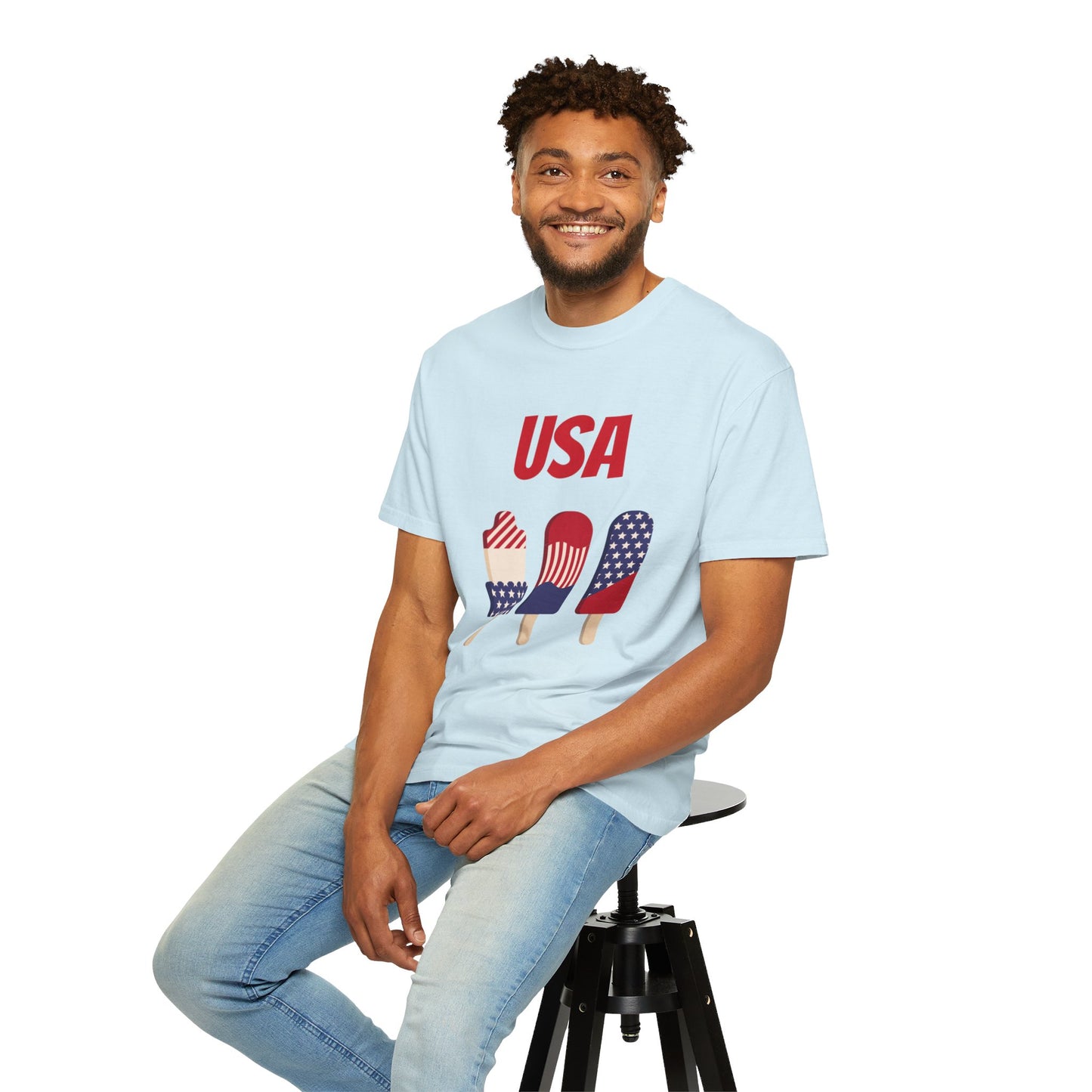 A man wearing a light blue Unisex USA T-shirt: 3 colors; Cotton; Comfort Colors 1717 with popsicle designs is sitting on a stool. Made from Printify's 100% ring-spun cotton, he is smiling and has a casual posture.