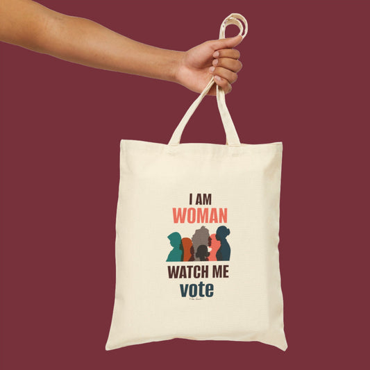 Hand holding a Printify Voting Womens Tote Bag: 15" x 16": Cotton Canvas; Graphics made of 100% cotton canvas with text "I AM WOMAN WATCH ME vote" and silhouettes of three women in different colors. Maroon background. USA assembled for quality you can trust.