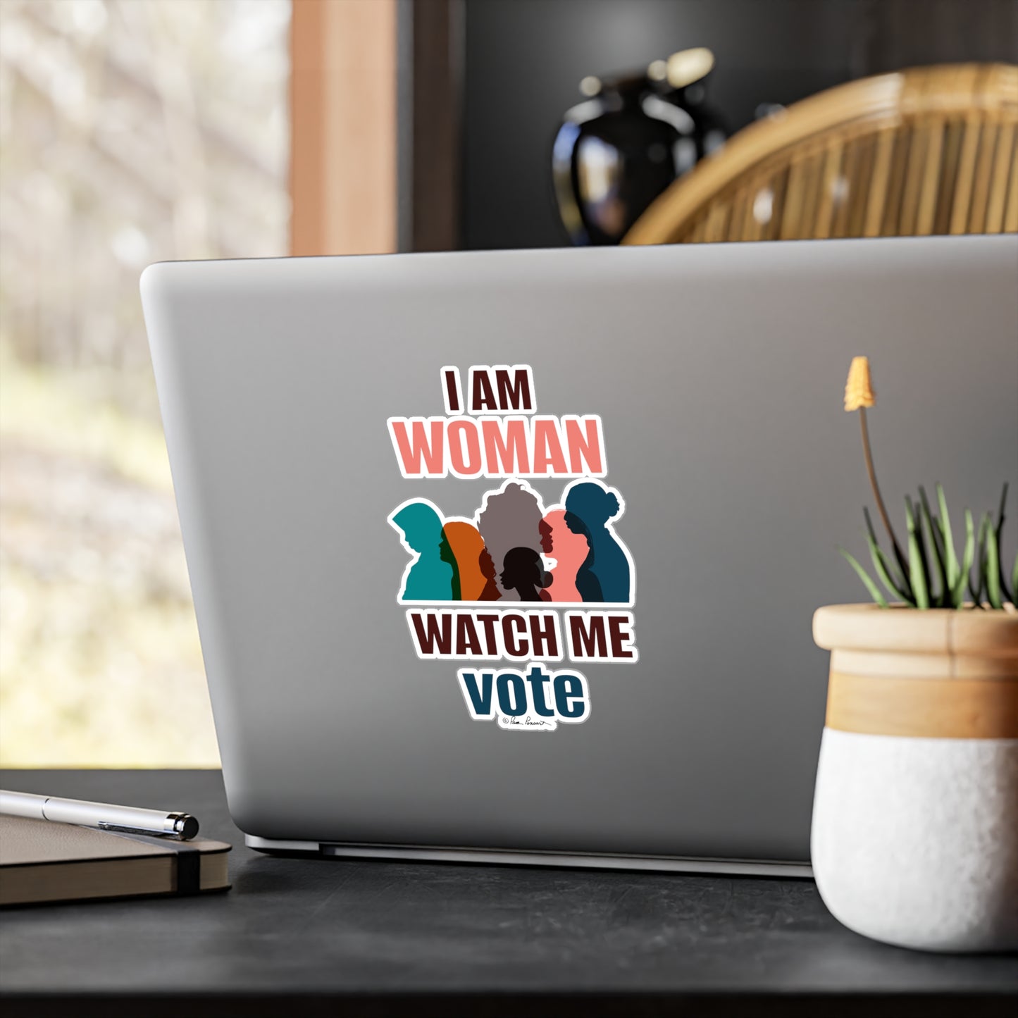 A laptop with a removable Printify Voting Women's Decal sticker reading "i am woman watch me vote" featuring silhouettes of four women, placed on a desk near a window.