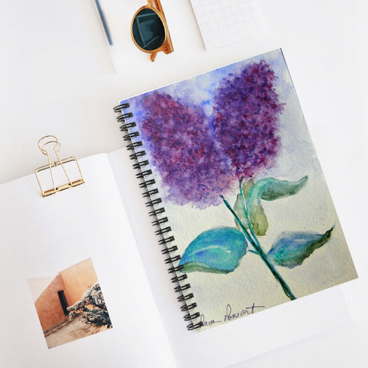 A Purple Lilacs Notebook by Printify, featuring watercolor art, rests elegantly on a white surface beside an open book, sunglasses, and a notepad.