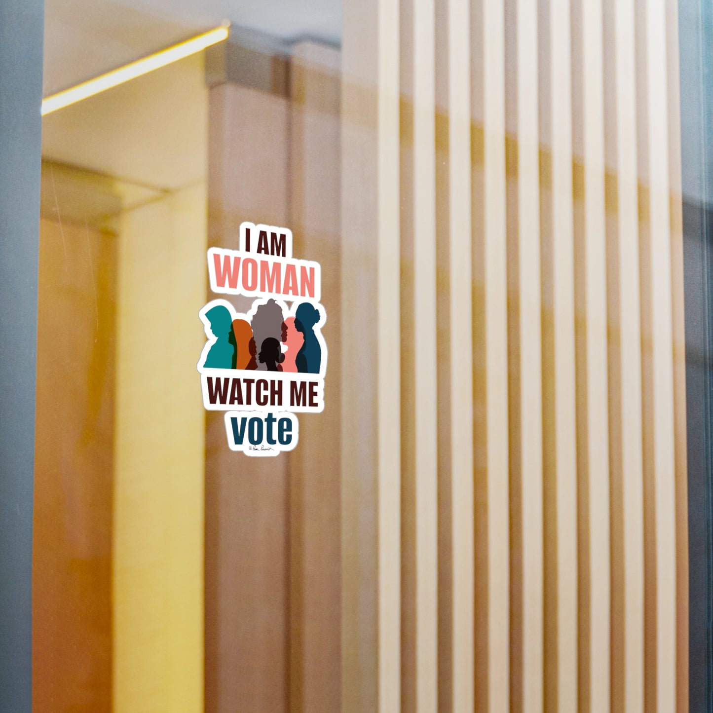 A Printify removable adhesive sticker on a glass window that reads "i am woman watch me vote," featuring silhouettes of three women, with a blurred background of vertical blinds.