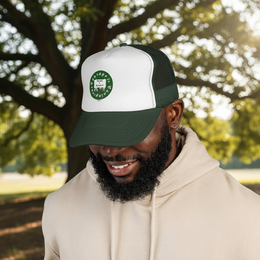 A man stands outdoors in front of a large tree wearing a beige hoodie and the Printify Courage is Contagious Trucker-Cap (Unisex, 2 colors) with a green and white mesh back and circular logo.