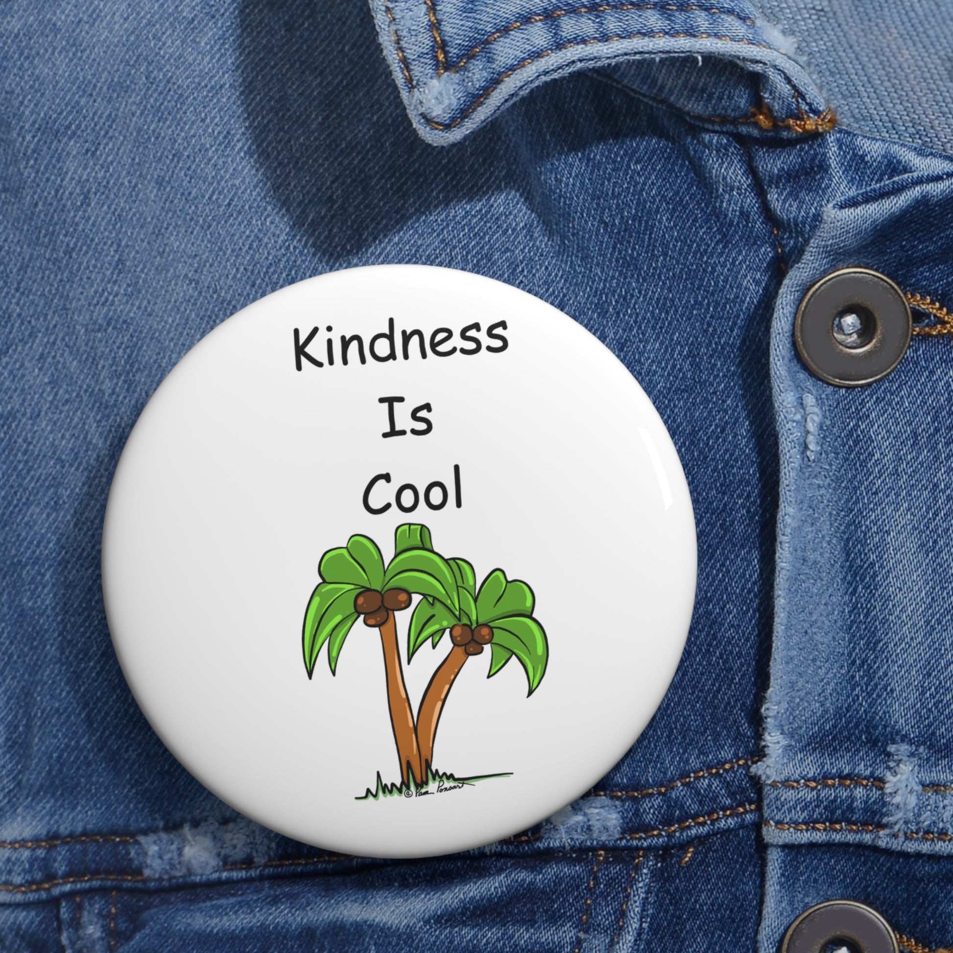 A Printify Unisex Kindness-Is-Cool Button, made of lightweight metal and available in 2 sizes, is pinned to a denim jacket. It features the text "Kindness Is Cool" along with an illustration of two palm trees and includes a safety pin backing for easy attachment.