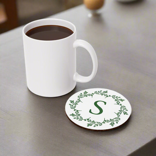 A white mug filled with black coffee sits on a gray surface beside a Printify Custom Letter Coaster: 2 styles; Cork-back; Personalized featuring a green letter 'S' surrounded by a leafy design.