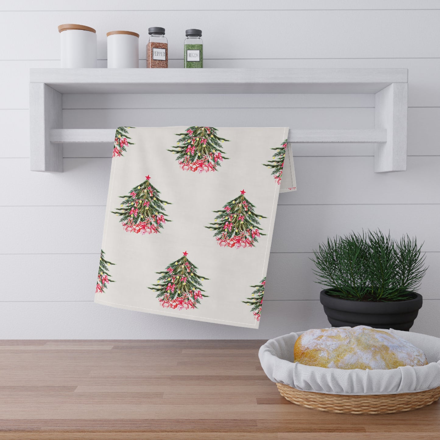 A cheerful kitchen display features Printify's Christmas-Tree Kitchen Towels, measuring 18" x 30", draped on a rack and embodying the essence of the holiday season. Spices line a shelf, a potted plant enhances the countertop, and a basket with cloth graces the wooden surface, all brought to life with vivid colors.