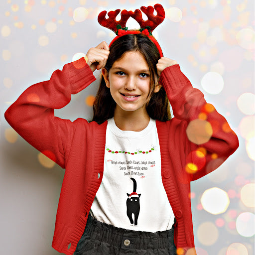 A young person wearing a red sweater and reindeer antlers smiles, revealing a Printify Festive Kids Holiday T-shirt featuring Santa Claws. The unisex Gildan shirt, crafted from ethically sourced cotton, peeks through beneath the sweater. The background shimmers with circular light bokeh.