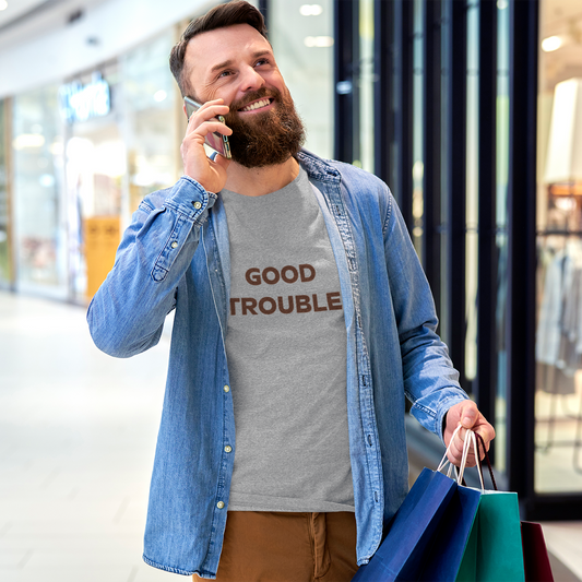 A bearded man smiles while talking on the phone and holding shopping bags in a mall. He wears a grey Printify Good Trouble Unisex T-shirt by Gildan, a stylish cotton shirt available in 3 colors.