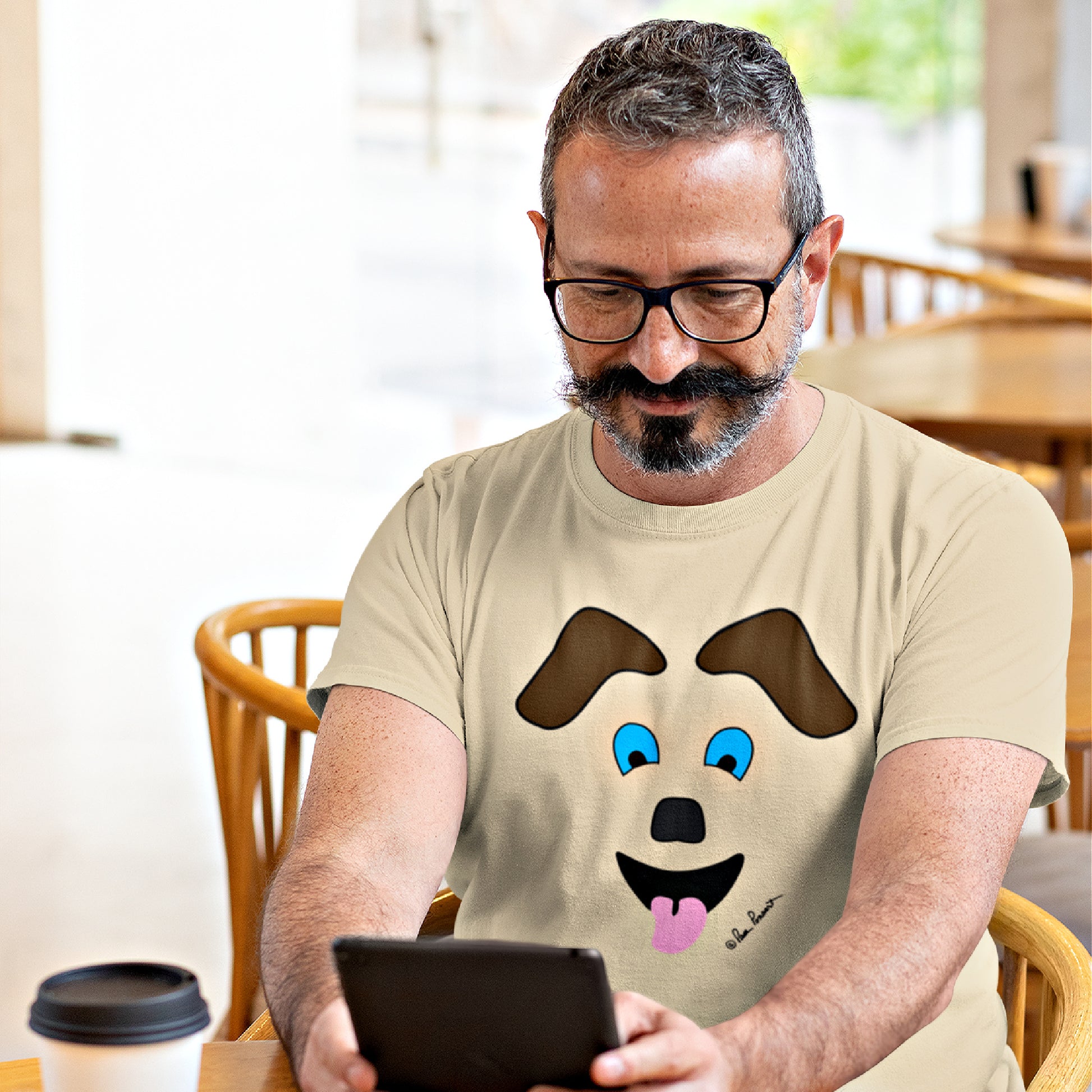 Mock up of a man reading his tablet in a café while wearing the Sand-colored t-shirt