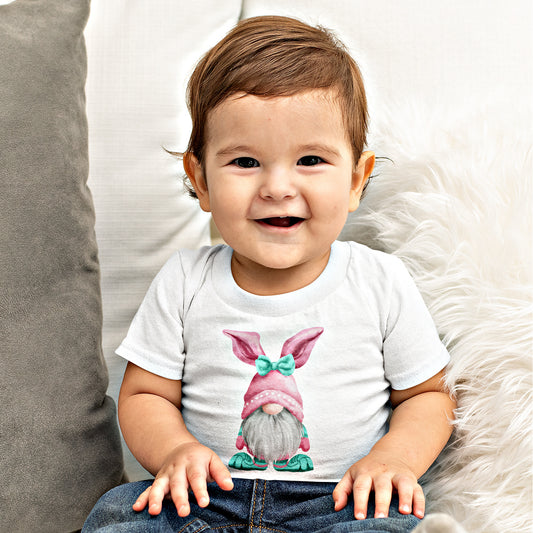 A happy toddler wearing a Printify Baby's Easter-Gnome T-shirt sits on a cozy surface.