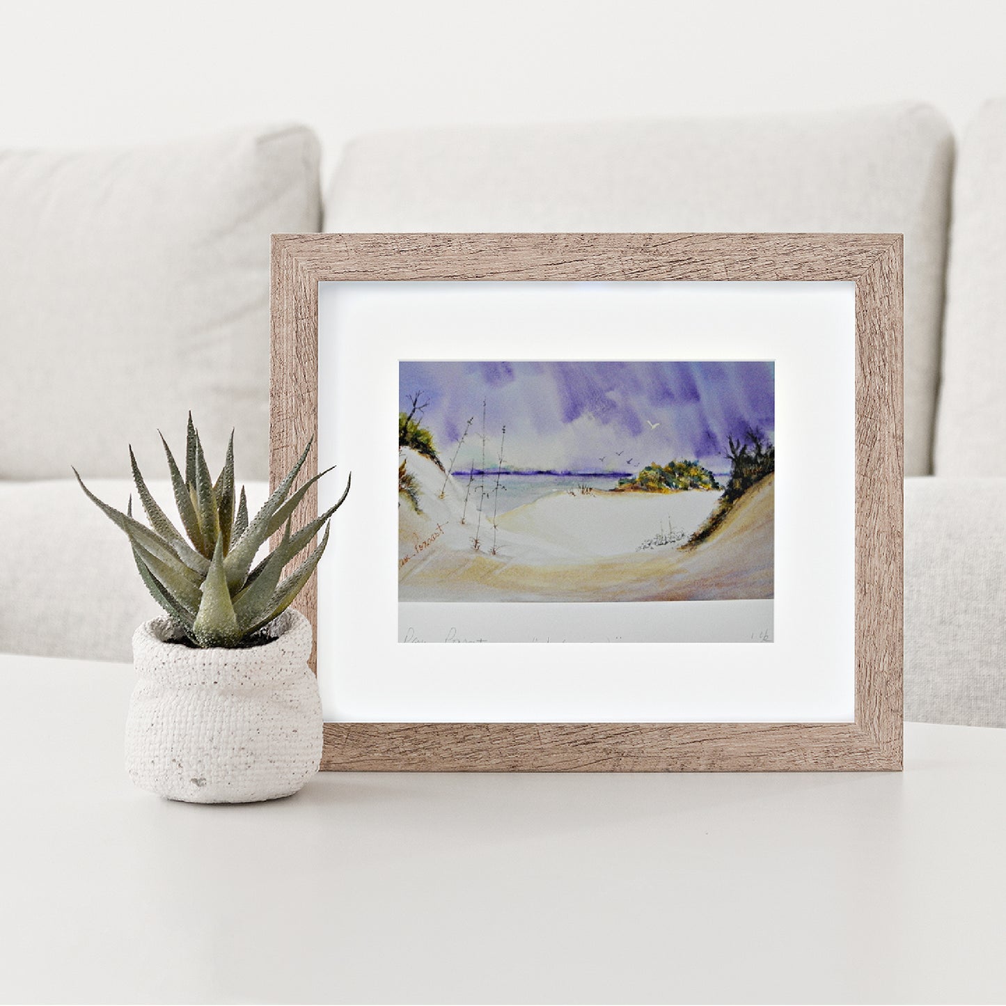 Mock up of our Beach Décor Print within a wood frame with white mat on a table with a potted aloe plant on the left hand side and pillows in the background.