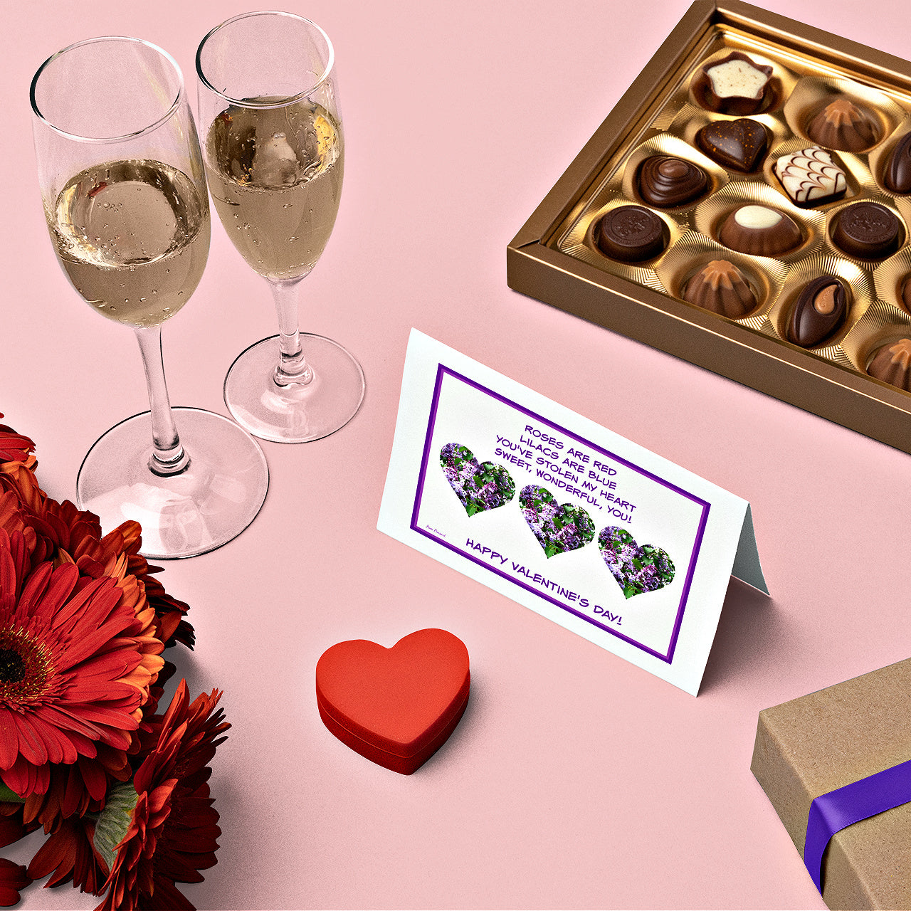Mock up of the card surrounded by a box of chocolates, red flowers and glasses of a bubbly beverage