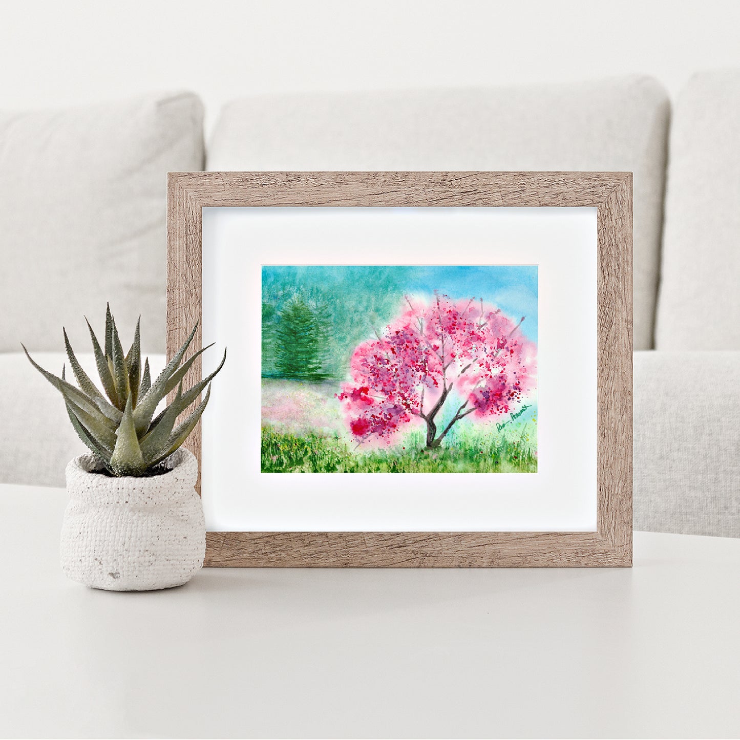 Mock up of our Pink Watercolor Print depicting a pink tree in full bloom in a meadow surrounded by a white mat and a brown wood frame (frame not included)