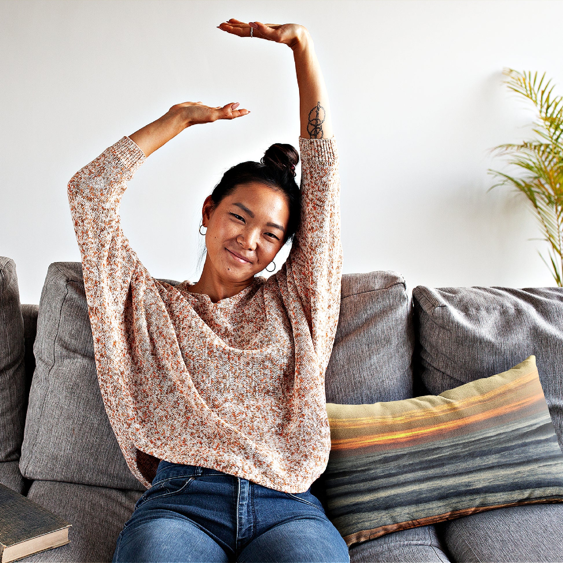 Mock up of a smiling girl, stretching her arms while sitting on a sofa with our Orange-Sunset Lumbar Pillow positioned on her left side