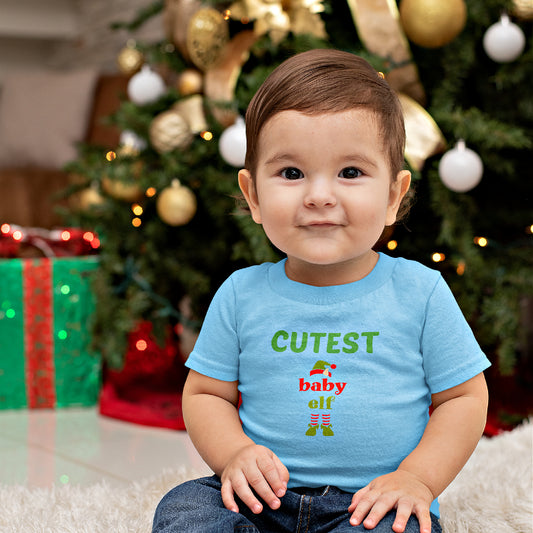 Mock up of a baby boy sitting by a Christmas tree while wearing our blue Baby Elf T-shirt