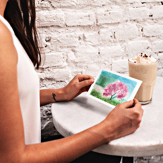 Girl looking at the front of our Watercolor Landscape Card while sitting down at a white café table