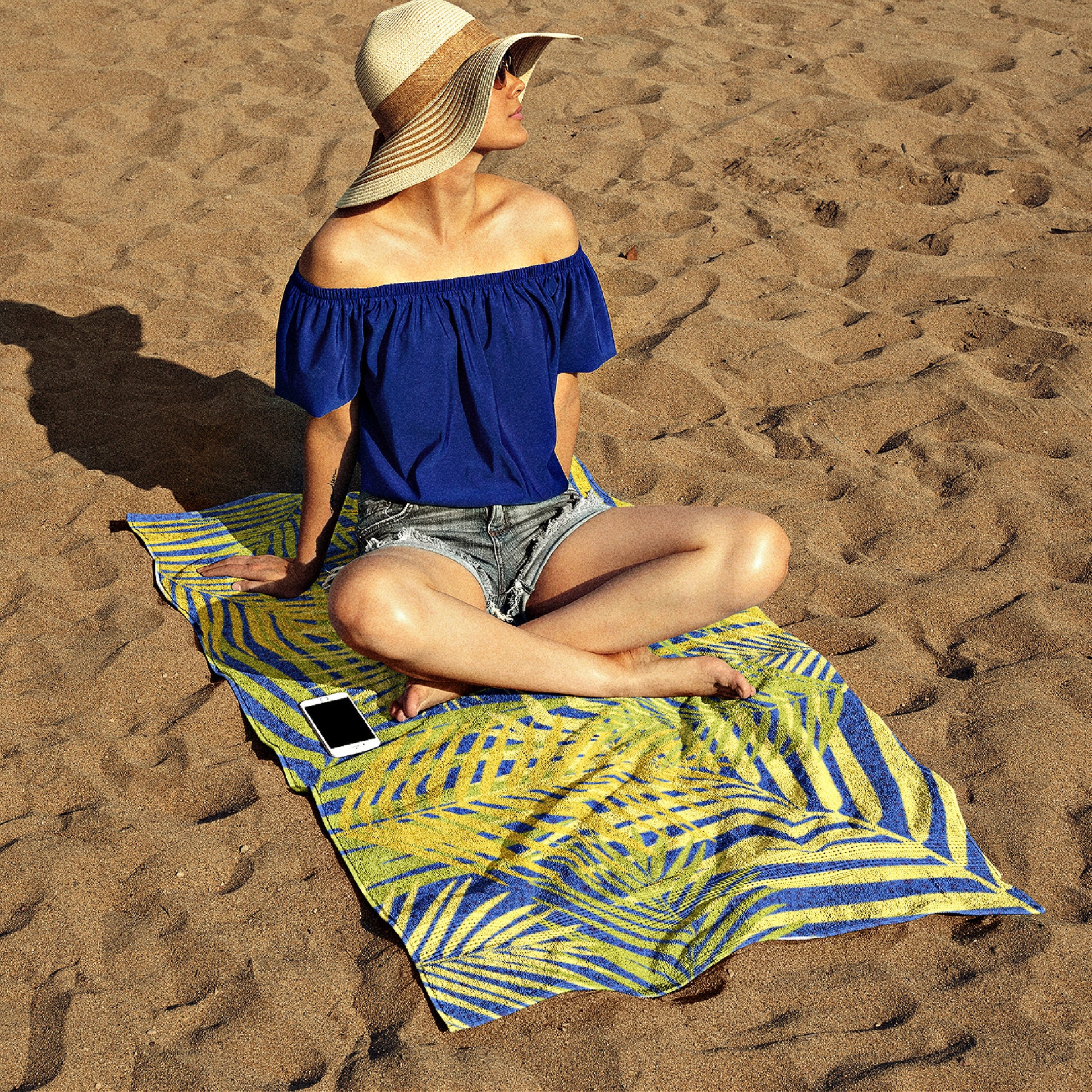 Mock up of a young lady, wearing a straw hat, is sitting our our poly-cotton beach towel